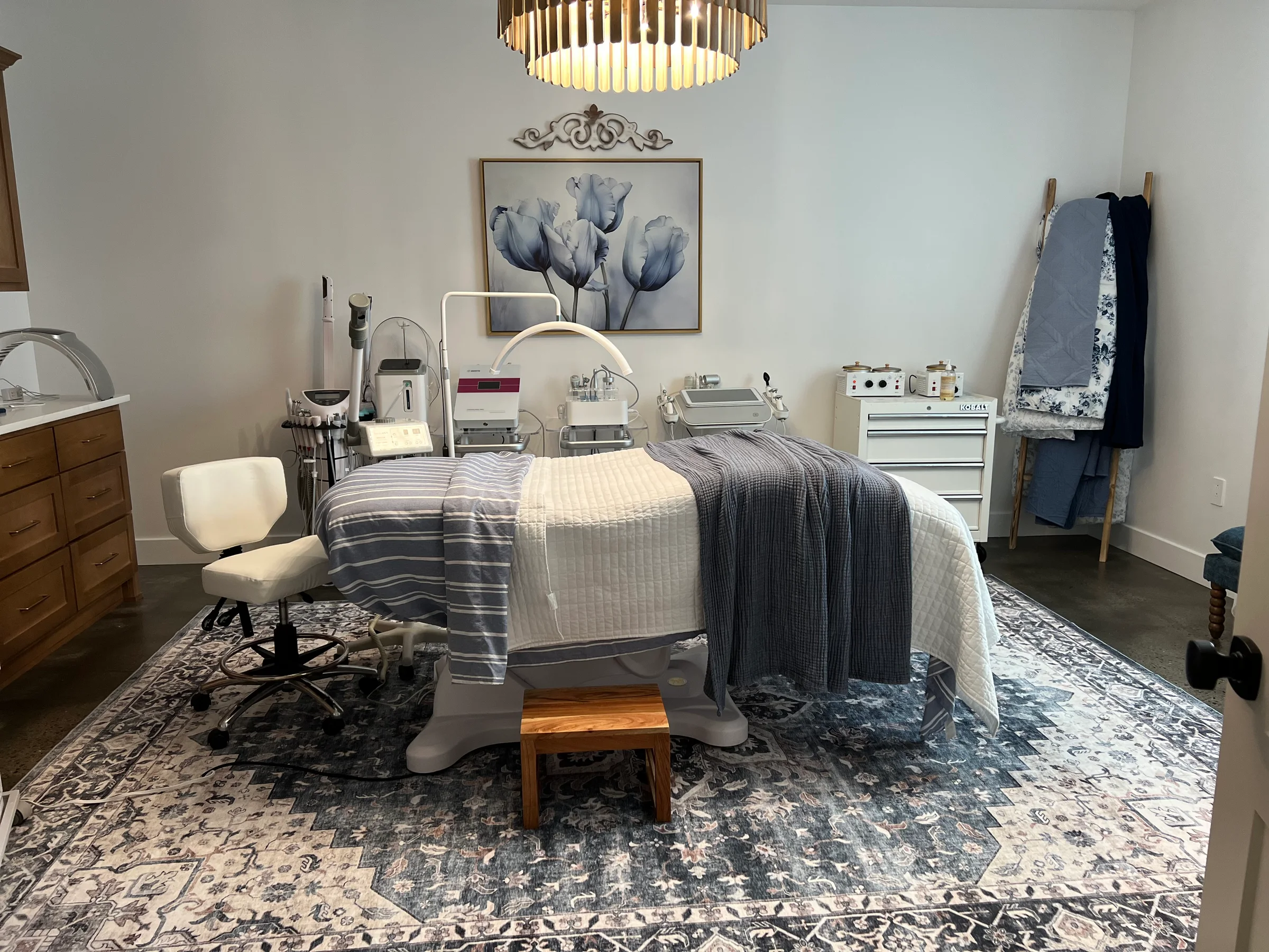 The treatment room at Southern Ground Esthetics with a facial bed layered in soft grey linens, professional skincare equipment, a gold chandelier, and a blue patterned rug