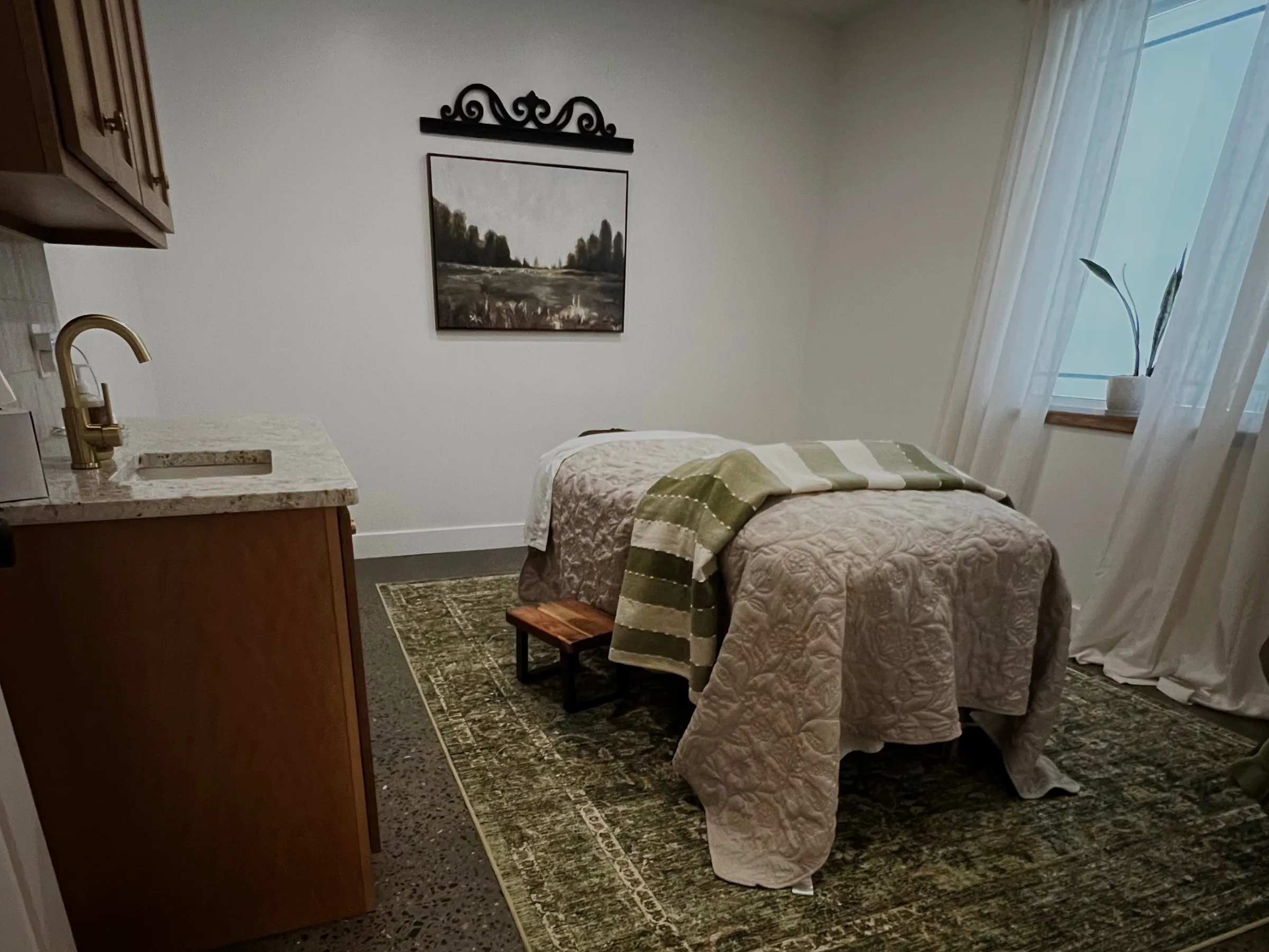 The massage room at Southern Ground Esthetics with a massage table dressed in a cream quilted coverlet and green striped throw, a granite-topped wood vanity with gold fixtures, a landscape painting, and a green vintage rug over polished concrete